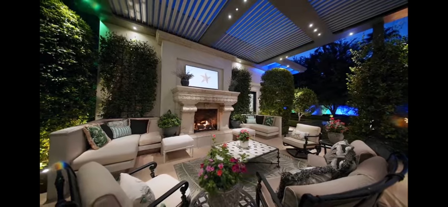 Covered estate patio with fireplace, manicured greenery, and evening lighting